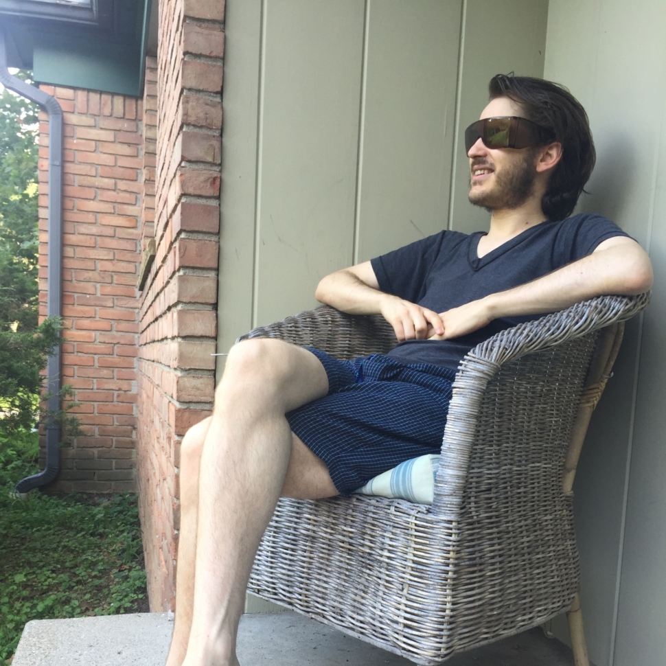 Image of Glenn, a white male with black hair and beard, sitting outside in a wicker chair. He is wearing wrap-around sunglasses