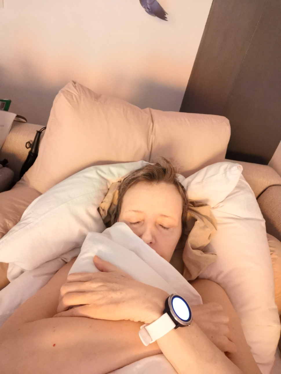 Picture of Nicole, a white woman with short light brown hair. She is lying down with eyes closed, holding a pillow. She is wearing a medical alert smart watch
